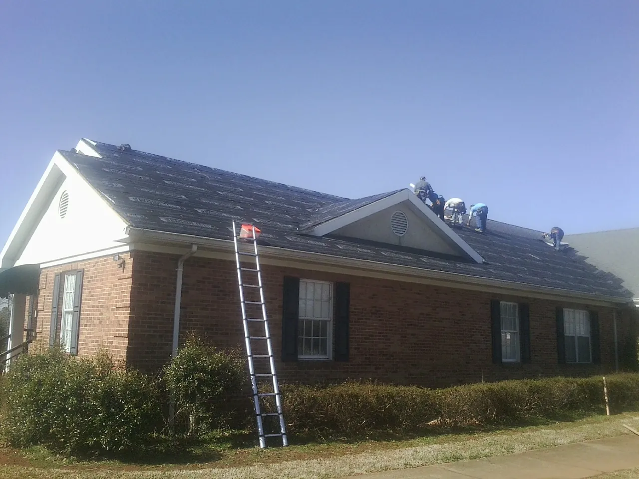 Crew tearing shingles off a brick ranch roof with a ladder set — Emergency Roof Repair work by our crew in Williston