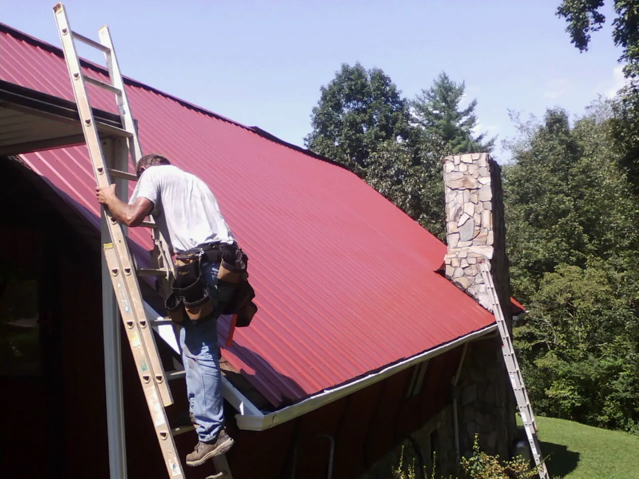 Roofer on a ladder beside a red metal roof and stone chimney — Soffit & Fascia Repair by Roofing Brothers in Williston